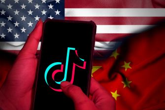 A hand holding a smartphone displaying the TikTok logo, set against the backgrounds of the American and Chinese flags.