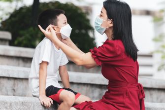 A woman helps a young boy put on a protective face mask outdoors.