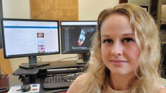 A woman with wavy blonde hair sits at her desk with two computer monitors displaying data visualizations and information related to COVID-19 statistics.