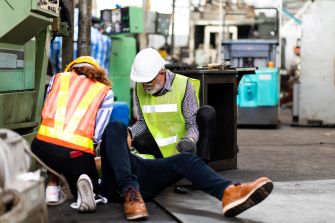 A workplace accident scene with two safety personnel attending to an injured worker on the floor.