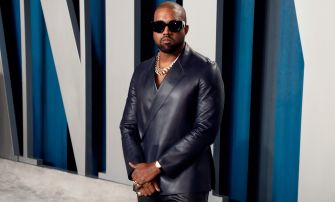 A man wearing sunglasses and a stylish black outfit poses confidently, with a serious expression, against a modern, blue-patterned background.