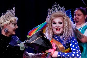 Drag performers celebrating during a performance, with one holding flowers and wearing a colorful outfit.