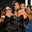 Lizzo speaking at an awards event, wearing a black dress and holding a microphone, surrounded by her team. Lizzo speaking at an awards event, wearing a black dress and holding a microphone, surrounded by her team.