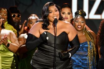Lizzo speaking at an awards event, wearing a black dress and holding a microphone, surrounded by her team.