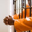 A person in an orange prison jumpsuit resting their hands on prison bars. A person in an orange prison jumpsuit resting their hands on prison bars.