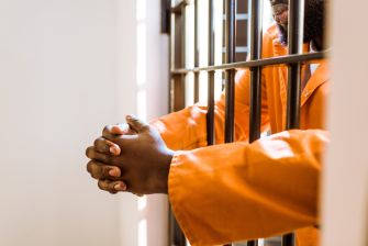 A person in an orange prison jumpsuit resting their hands on prison bars.