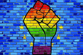 A raised fist symbol with a rainbow gradient against a blue brick wall, representing LGBTQ+ rights and activism.