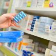 A person holding a blister pack of medication in a pharmacy, with various pharmaceutical boxes in the background. A person holding a blister pack of medication in a pharmacy, with various pharmaceutical boxes in the background.