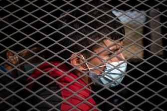 A young boy wearing a mask is seen behind a metal grid, highlighting the conditions faced by unaccompanied minors at the U.S. southern border.