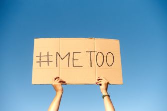 A person holding a cardboard sign that reads "#Me Too" against a blue sky.
