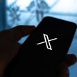 A person holding a smartphone displaying the X app interface on the screen. A person holding a smartphone displaying the X app interface on the screen.