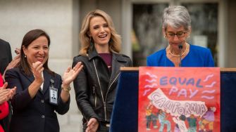 A group of people at a public event, with one woman speaking at a podium displaying a sign that reads "EQUAL PAY CALIFORNIA."