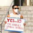 A person wearing a mask holds a sign that says "YES on 15 FUND SCHOOLS + PUBLIC SERVICES" while standing on steps. A person wearing a mask holds a sign that says "YES on 15 FUND SCHOOLS + PUBLIC SERVICES" while standing on steps.