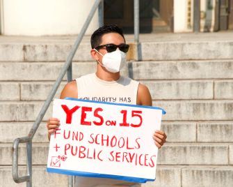 A person wearing a mask holds a sign that says "YES on 15 FUND SCHOOLS + PUBLIC SERVICES" while standing on steps.