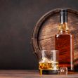 A bottle of whiskey next to a glass with ice, set against a background featuring a wooden barrel. A bottle of whiskey next to a glass with ice, set against a background featuring a wooden barrel.