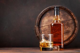 A bottle of whiskey next to a glass with ice, set against a background featuring a wooden barrel.