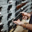 Person examining a handgun in a firearm retailer, showcasing various handguns displayed on a wall. Person examining a handgun in a firearm retailer, showcasing various handguns displayed on a wall.