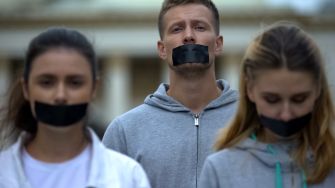 Three individuals with tape over their mouths, symbolizing silenced voices, stand together outdoors.