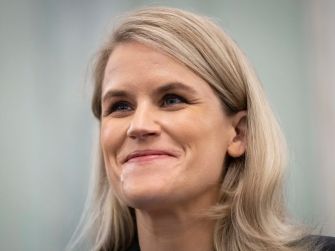A close-up of a smiling woman with long, blonde hair, seated in a soft-focus setting.