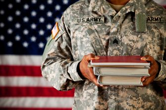 A military student in uniform holds books in front of an American flag, symbolizing his commitment to education and service.