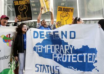 Protesters holding signs and a large banner advocating for the defense of Temporary Protected Status (TPS) for Hondurans and Nicaraguans.