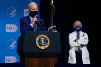 President Biden speaking at a podium with the presidential seal, with Dr. Anthony Fauci visible in the background, both wearing masks.