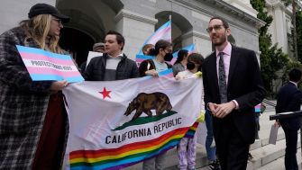 A group of LGBTQ activists holding a California state flag with a rainbow stripe and signs advocating for LGBTQ rights outside a government building.