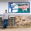 A farmer standing in front of the Faust Farms sign, holding a cane at his dairy farm. A farmer standing in front of the Faust Farms sign, holding a cane at his dairy farm.