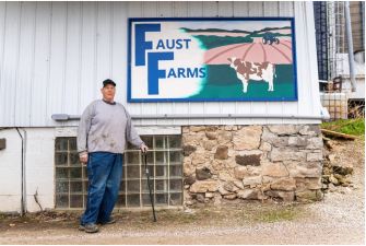 A farmer standing in front of the Faust Farms sign, holding a cane at his dairy farm.