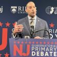 A man speaking at a gubernatorial primary debate podium with a background featuring political branding and logos. A man speaking at a gubernatorial primary debate podium with a background featuring political branding and logos.