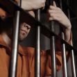 A close-up of a person in an orange prison jumpsuit holding onto metal bars. A close-up of a person in an orange prison jumpsuit holding onto metal bars.