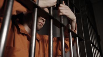 A close-up of a person in an orange prison jumpsuit holding onto metal bars.