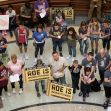 A group of people gathers inside a building, holding signs related to abortion rights, with some signs stating "Roe is unconstitutional" and "We will adopt your baby." A group of people gathers inside a building, holding signs related to abortion rights, with some signs stating "Roe is unconstitutional" and "We will adopt your baby."
