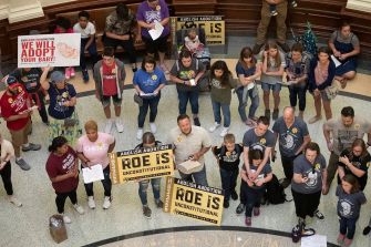 A group of people gathers inside a building, holding signs related to abortion rights, with some signs stating "Roe is unconstitutional" and "We will adopt your baby."