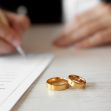 Image of two gold wedding rings on a table with a person writing on a document in the background. Image of two gold wedding rings on a table with a person writing on a document in the background.