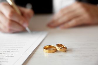Image of two gold wedding rings on a table with a person writing on a document in the background.