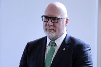 A man in a suit and glasses looks serious in a well-lit indoor setting.
