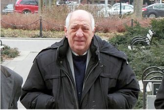 Elderly man in a black coat walking in a park setting, surrounded by trees and city buildings.