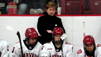 A woman stands behind hockey players on the bench during a game, appearing concerned or contemplative.