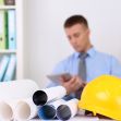 A yellow hard hat placed next to rolled blueprints, with a man in a suit looking at a tablet in the background. A yellow hard hat placed next to rolled blueprints, with a man in a suit looking at a tablet in the background.