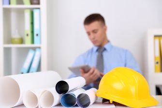 A yellow hard hat placed next to rolled blueprints, with a man in a suit looking at a tablet in the background.