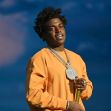 Rapper Kodak Black standing with a chain and microphone, wearing an orange sweatshirt. Rapper Kodak Black standing with a chain and microphone, wearing an orange sweatshirt.