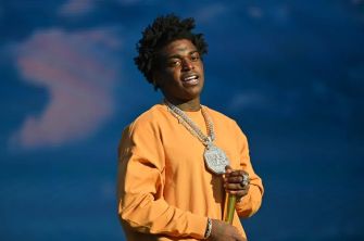 Rapper Kodak Black standing with a chain and microphone, wearing an orange sweatshirt.