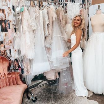 A smiling woman in a white dress stands next to a rack of wedding gowns in a bright studio setting.