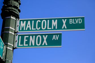Street signs for Malcolm X Boulevard and Lenox Avenue against a clear blue sky.