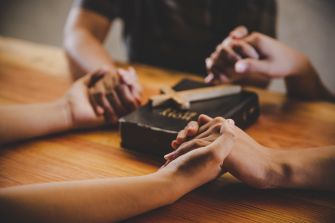 Hands clasped in prayer over a Bible on a wooden table.