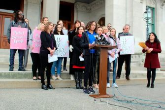 A group of athletes and supporters protesting for women's sports rights outside a government building, holding signs advocating for fair play and the protection of women's sports.