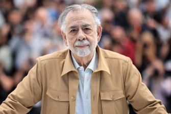 Francis Ford Coppola poses for a photo at a public event, wearing a beige jacket and standing in front of a blurred crowd.