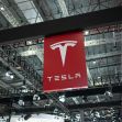 Logo of Tesla displayed at an automotive exhibition, surrounded by lights and structures. Logo of Tesla displayed at an automotive exhibition, surrounded by lights and structures.
