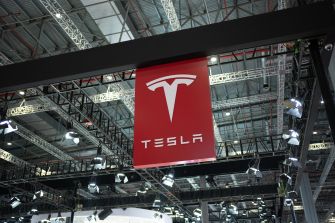 Logo of Tesla displayed at an automotive exhibition, surrounded by lights and structures.